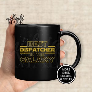 May include: Black coffee mug with a white design that says "Best Dispatcher in the Galaxy". The text is in a yellow font. The mug has a white handle.