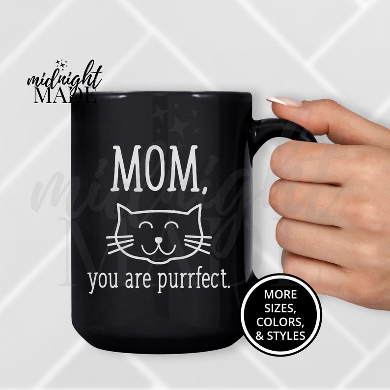 May include: Black ceramic coffee mug with white text that reads "MOM. you are purrfect." and a cartoon cat face. The mug has a white handle.