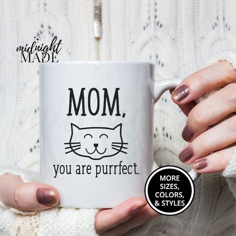 May include: White ceramic mug with black text that reads "MOM, you are purrfect."  A black cat face with closed eyes and a smile is above the text.  The mug is being held by a person's hand.  A black circle with white text reads "MORE SIZES, COLORS, & STYLES".