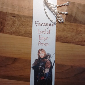 Könnte beinhalten: Ein weißes Lesezeichen mit einem Bild von Faramir, einer Figur aus Herr der Ringe, der einen Bogen und Pfeil hält. Das Lesezeichen trägt den Text "Faramir, Herr von Emyn Arnen" in brauner Tinte.