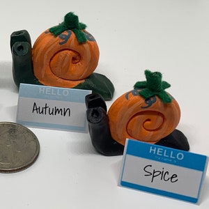May include: Four small pumpkin snail figurines with green leaves and black, purple, and green shells. Each snail has a name tag with the text "HELLO" and a name: "Munkin-My", "Pie", "Autumn", and "Spice".