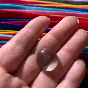 May include: A clear glass marble, slightly oval shaped, with a slight shimmer, held in a hand.