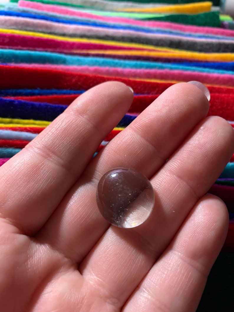 May include: A clear glass marble, slightly oval shaped, with a slight shimmer, held in a hand.