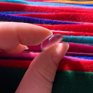 May include: A clear glass bead, held between two fingers, against a colorful striped background.