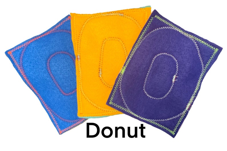 May include: Three felt squares with a donut shape cut out. The squares are blue, orange, and purple. The donut shapes are outlined in pink, yellow, and white thread.