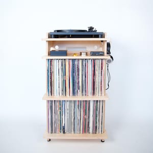 May include: A light wood record player stand with three shelves. The top shelf has a turntable and a black box. The middle and bottom shelves are filled with vinyl records.