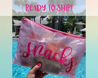 Puede incluir: Una bolsa de malla rosa con cremallera con la palabra "Snacks" impresa en rosa. La bolsa está llena de varios bocadillos y la está sujetando una mano cerca de una piscina.