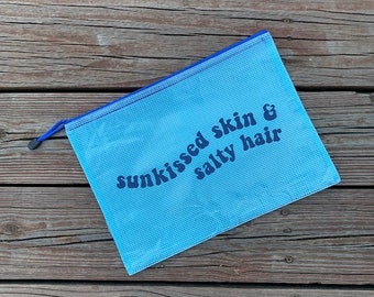 May include: A light blue mesh zippered pouch with a blue zipper pull. The pouch has the text "sunkissed skin & salty hair" printed in dark blue.