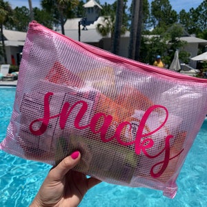 May include: A pink mesh bag with the word "snacks" printed in pink letters. The bag is filled with various snacks and is being held by a hand.