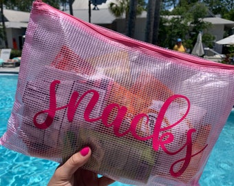 May include: A pink mesh bag with the word "snacks" printed in pink letters. The bag is filled with various snacks and is being held by a hand.