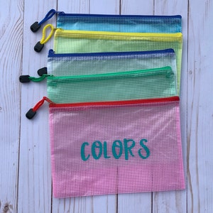 Puede incluir: Cinco bolsas de malla con cremallera en diferentes colores, apiladas sobre una superficie de madera blanca. La bolsa inferior es rosa y tiene la palabra "COLORS" escrita en turquesa en la parte delantera.