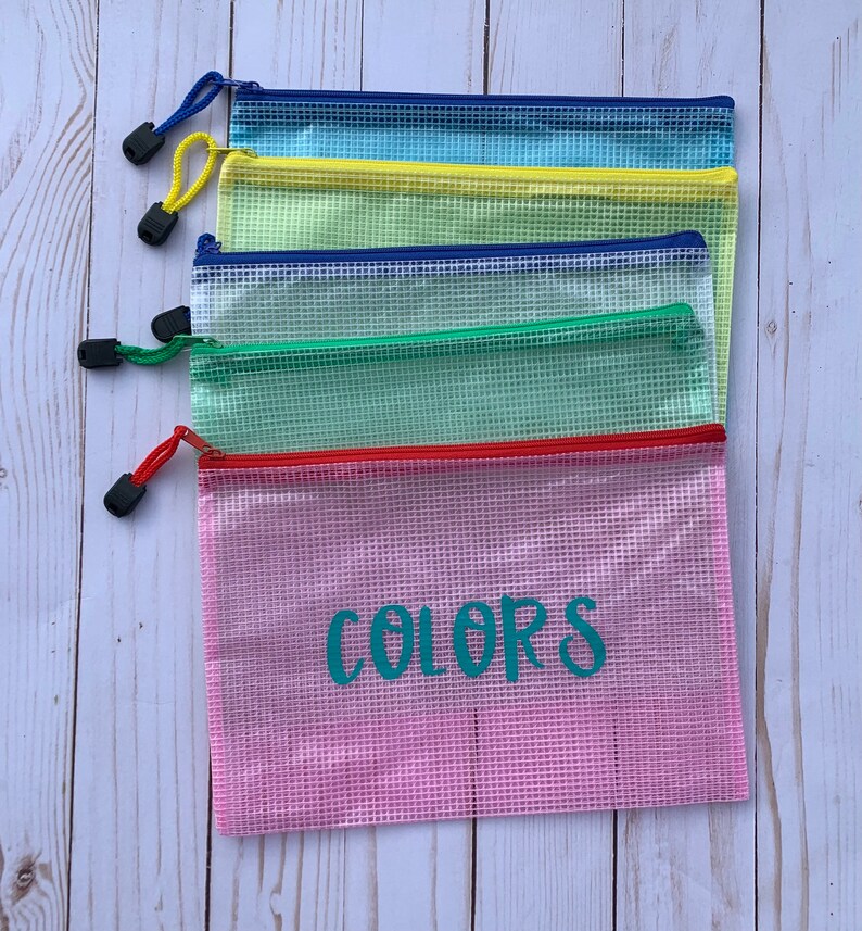 Puede incluir: Cinco bolsas de malla con cremallera en diferentes colores, apiladas sobre una superficie de madera blanca. La bolsa inferior es rosa y tiene la palabra "COLORS" escrita en turquesa en la parte delantera.