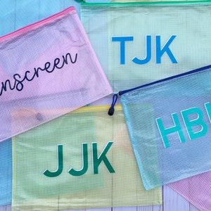 May include: Five mesh zippered pouches with different colored backgrounds and personalized text. The pouches are light blue, yellow, light blue, pink, and pink. The text on the pouches reads "Sunscreen", "TJK", "HBK", "JJK", and "Sunscreen".