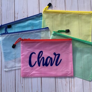 Puede incluir: Cinco bolsas de malla con cremallera en diferentes colores, incluyendo azul, amarillo, rosa y verde. La bolsa rosa tiene el nombre "Char" escrito en azul.