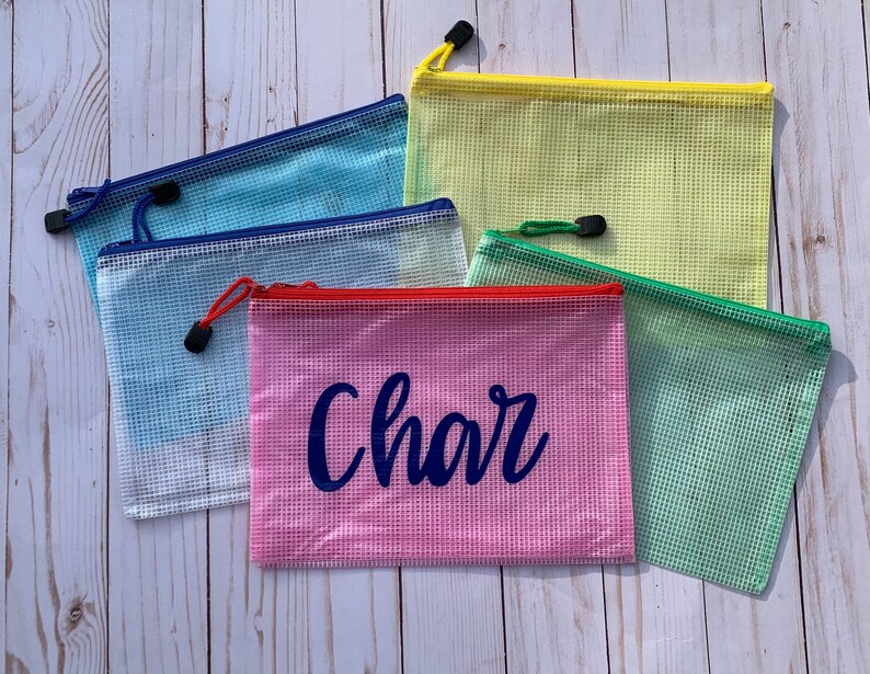 Puede incluir: Cinco bolsas de malla con cremallera en diferentes colores, incluyendo azul, amarillo, rosa y verde. La bolsa rosa tiene el nombre "Char" escrito en azul.