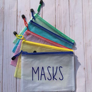 Puede incluir: Seis bolsas de malla con cremallera de colores con tiradores azules, apiladas unas sobre otras. La bolsa inferior es blanca con la palabra "MASKS" impresa en azul.