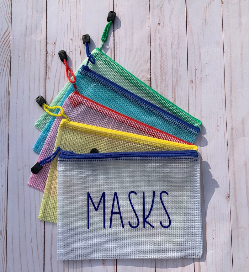 Puede incluir: Seis bolsas de malla con cremallera de colores con tiradores azules, apiladas unas sobre otras. La bolsa inferior es blanca con la palabra "MASKS" impresa en azul.