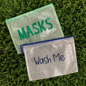 Puede incluir: Dos bolsas de pl&aacute;stico transparente con cremallera y tejido de malla. Una bolsa es verde y dice "MASKS" en letras verdes. La otra bolsa es blanca y dice "Wash Me" en letras azules.