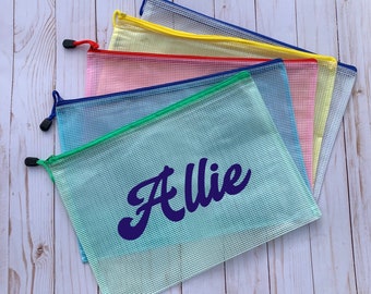 Puede incluir: Cinco bolsas de malla con cremallera de colores con una cremallera azul. La bolsa de la parte delantera es verde y tiene el nombre "Allie" escrito en morado.