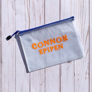 Puede incluir: Una bolsa de malla blanca con cremallera azul. La bolsa tiene las palabras "CONNOR EPIPEN" impresas en naranja.