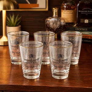 May include: A set of five clear glass tumblers with gold-toned rims, each featuring a black graphic of the Indianapolis Motor Speedway logo, a list of race winners, and the signature of Tony Hulman printed on the front of the glass surface.