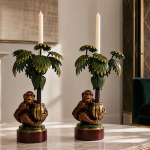May include: A pair of candle holders featuring brown monkey figures sitting on circular red bases. Each monkey holds a central stem topped with green palm-like leaves and a single white taper candle. The design incorporates gold accents.