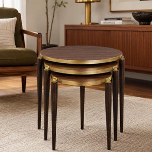 Puede incluir: Un juego de tres mesas nido con tableros circulares de madera oscura y bordes metálicos en tono dorado. Cada mesa se apoya sobre tres patas cónicas de madera oscura con detalles en tono dorado en la parte superior de las patas.