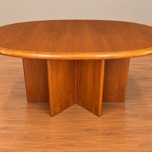 Peut inclure: Une table à manger ovale en bois avec une finition grainée brun clair lisse. La table présente un plateau épais et solide avec un bord arrondi, soutenu par un pied central composé de quatre panneaux de bois verticaux entrecroisés.