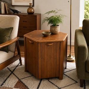 Op de afbeelding: Een zeshoekige houten bijzettafel met een gladde bruine afwerking, voorzien van een scharnierend blad en kastdeuren met geïntegreerde handgrepen. Op het tafelblad staan een kleine keramische kom en een groene varen in een beige pot.