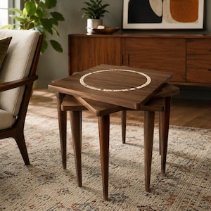 May include: A set of three nested square tables crafted from dark brown wood, featuring tapered legs and a circular inlay pattern on the top surface of the largest table. The tables are arranged in a staggered, overlapping formation.
