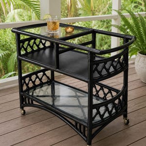 May include: A black bar cart featuring a bamboo-style frame with a lattice pattern on the sides. The cart has two tiers, each with a glass shelf, and is supported by four small metal casters. A glass of amber liquid with a lime slice sits on top.