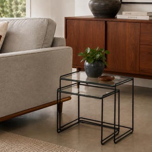 May include: A set of two nesting tables featuring clear glass rectangular tops and thin black metal frames. The tables are positioned together, with the smaller table partially tucked underneath the larger one, creating a tiered surface.