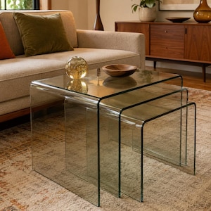 May include: A set of three clear glass nesting tables with a waterfall design. The tables are arranged in a cascading formation, featuring smooth, curved edges and a transparent construction that allows for a clear view of the floor beneath them.