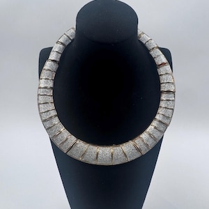 May include: A silver-toned necklace with a textured, geometric design. The necklace is displayed on a black mannequin head against a white background. The necklace has a gold-colored trim.