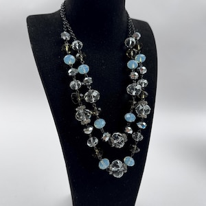 May include: A two-strand necklace with clear, light blue, and smoky gray faceted beads. The necklace is displayed on a black mannequin bust against a white background. The beads are strung on a dark chain.