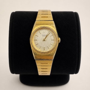 Può includere: Un orologio da polso Seiko Quartz color oro con quadrante argentato testurizzato, indici e lancette dorate sottili. L'orologio presenta un quadrante rotondo con una lunetta decorativa e un cinturino a maglie in metallo dorato abbinato.