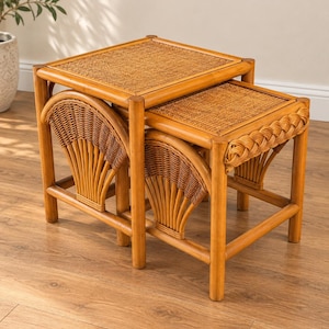 May include: A set of two nested tables constructed from light brown rattan. Each table features a square top with a woven cane surface and side panels designed with a fan-shaped wicker pattern. The legs are connected by horizontal support bars.