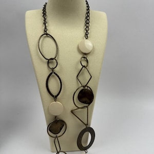 May include: A long necklace with a variety of shapes and colors. The necklace features a dark chain and bronze-colored metal rings, ovals, and triangles. It also includes cream-colored circular and oval accents. The necklace is displayed on a mannequin.