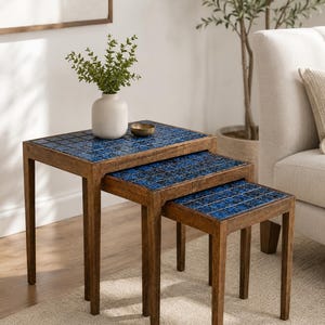 May include: A set of three nesting tables with dark brown wooden frames and square tabletops inlaid with small, glossy blue tiles. The tables are arranged in a cascading formation, each featuring four straight legs and a smooth, polished finish.