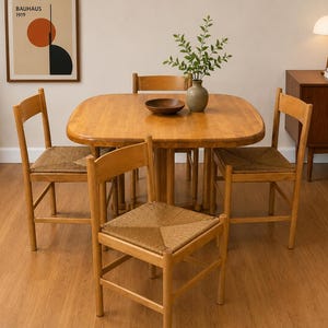 May include: A light brown wooden dining set consisting of a square table with rounded corners and four chairs. Each chair features a woven rush seat and a horizontal backrest. A small wooden bowl and a green plant in a vase sit on the table.