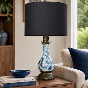 May include: A table lamp with a solid black cylindrical fabric shade and a decorative ceramic base featuring a blue and white collage of historical portraits and patterns, set upon an ornate, dark metallic pedestal with a brass-toned neck.