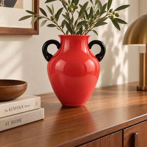 May include: A bright red ceramic vase with a glossy finish and two curved black handles on the sides. The vase has a wide, rounded body and a flared rim, containing a green leafy branch with small round olives extending from the top opening.