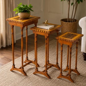 May include: A set of three graduated wooden nesting tables with square tops featuring a dark brown and tan striped border and a central patterned inlay. Each table stands on four slender legs connected by a cross-shaped base support.