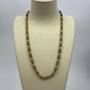 May include: A gold-toned necklace with a bamboo-like design. The necklace is displayed on a mannequin. The necklace features alternating sections of gold-colored beads and darker, cylindrical segments, creating a textured, natural look. The necklace is a jewelry item.