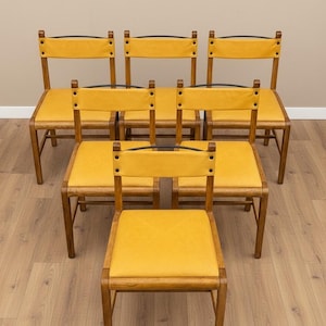 May include: A set of five wooden chairs with mustard yellow upholstered seats and backrests. Each chair features a dark metal curved bar across the top of the backrest, secured with visible rivets on the sides of the yellow upholstery panels.