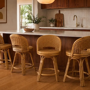 May include: Four light brown rattan bar stools with rounded backs and swivel seats. Each stool features a woven cane pattern on the backrest and a circular footrest connecting the four legs, providing a stable base for seating at a counter.