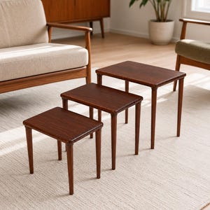 Puede incluir: Un juego de tres mesas nido de madera de color marrón oscuro, cada una con una superficie rectangular de esquinas redondeadas y cuatro patas delgadas y cónicas. Las mesas están dispuestas en forma escalonada, mostrando alturas variables.