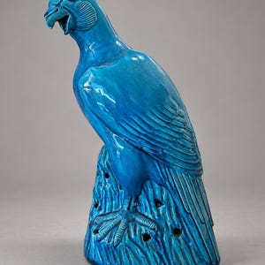 May include: A glossy, bright turquoise ceramic sculpture of a bird of prey perched on a textured, stump-like base. The bird is depicted with its head turned, beak slightly open, and detailed wing feathers, featuring small circular holes in the base.