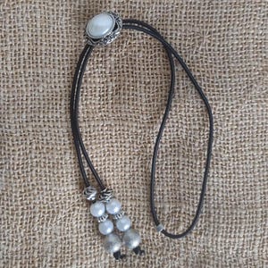 May include: A bolo tie featuring a silver-toned oval pendant with a white stone center, surrounded by a decorative border. The black cord is adorned with white and silver beads, and silver-toned decorative accents.