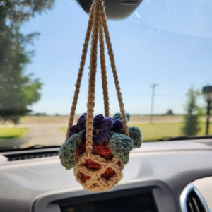 May include: A crocheted hanging plant with purple flowers and green leaves, hanging from the rear view mirror of a car.